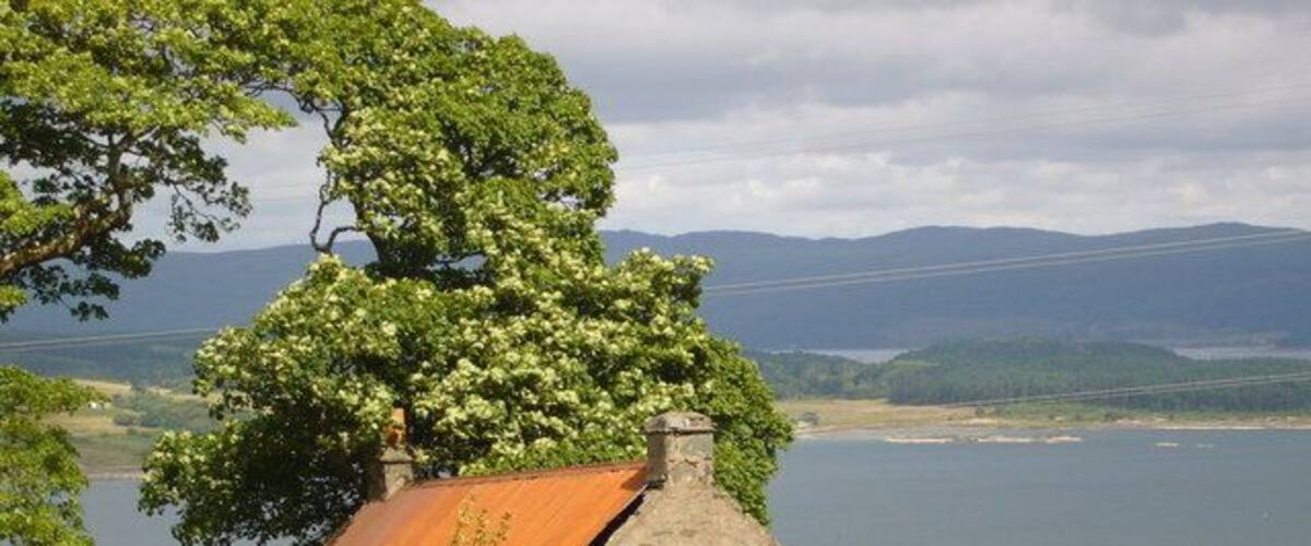 Una casa de piedra con techo oxidado, rodeada de hierba y un gran árbol, con vista a un lago y colinas lejanas.