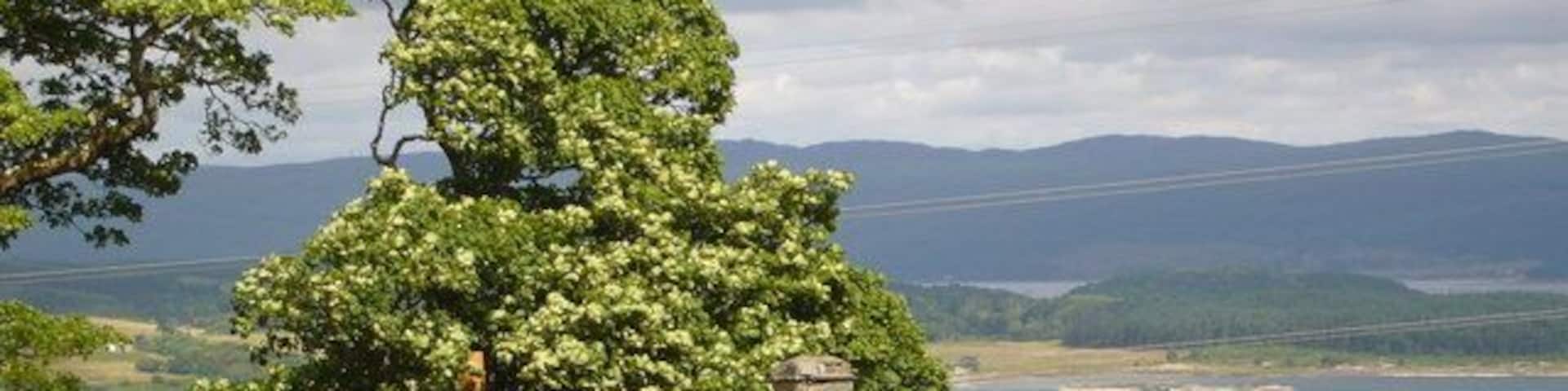 A stone house with a rusted roof, surrounded by grass and a large tree, overlooking a lake and distant hills.