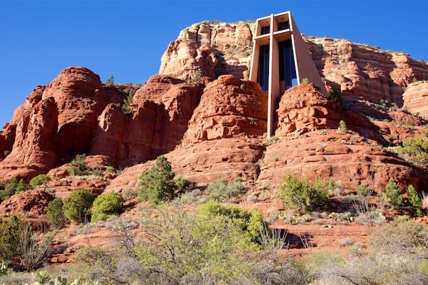 Formación rocosa roja con una cruz en la cima y un edificio con ventanas amplias.