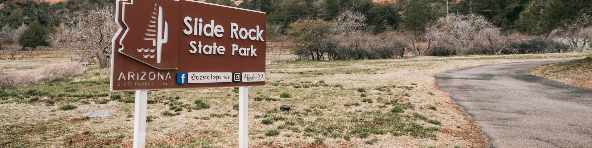 A sign for Slide Rock State Park in Arizona, with a road curving to the right and red rock formations in the background.
