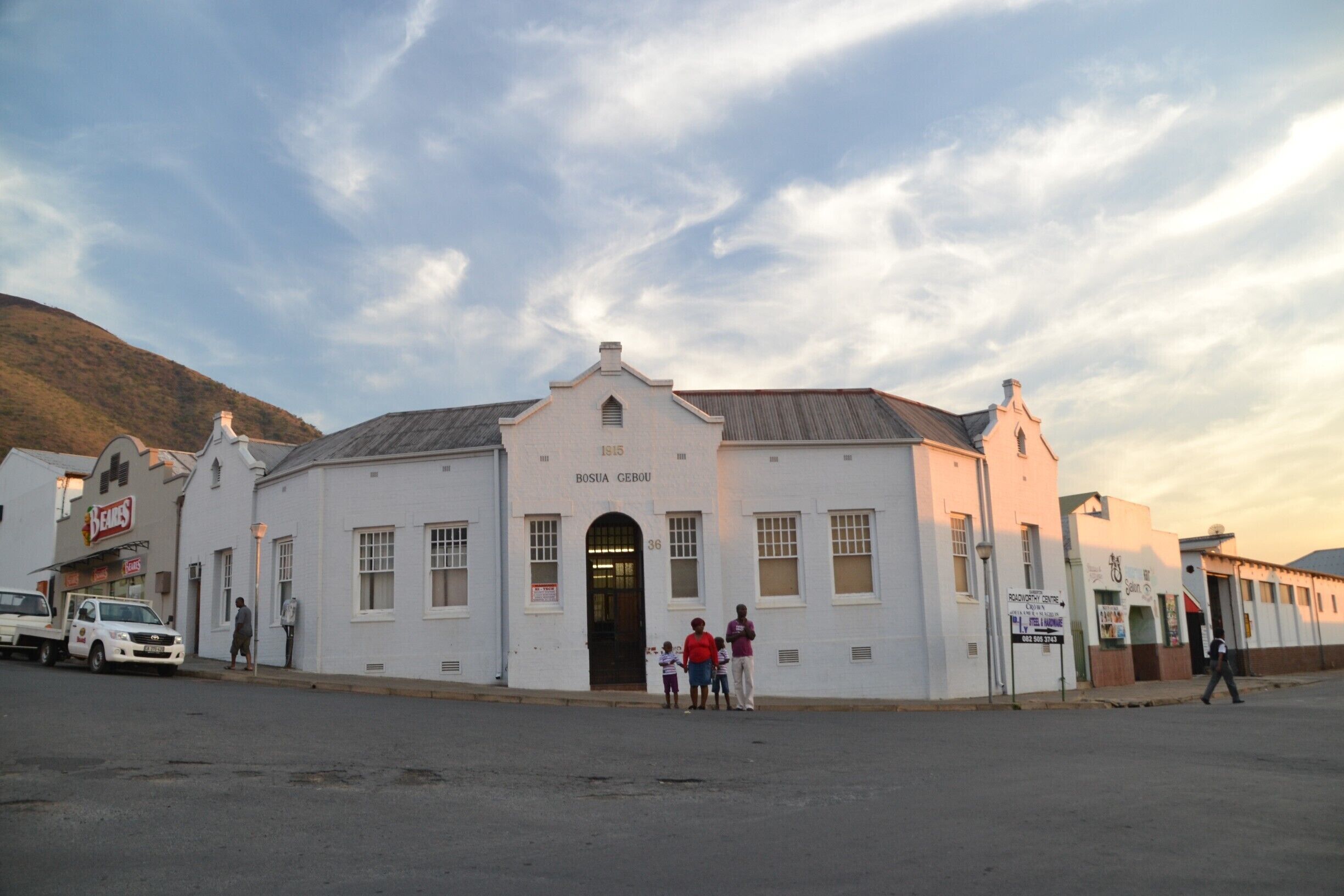 A white building with a sign reading "BOSIA GEBOU" and a few people standing in front.