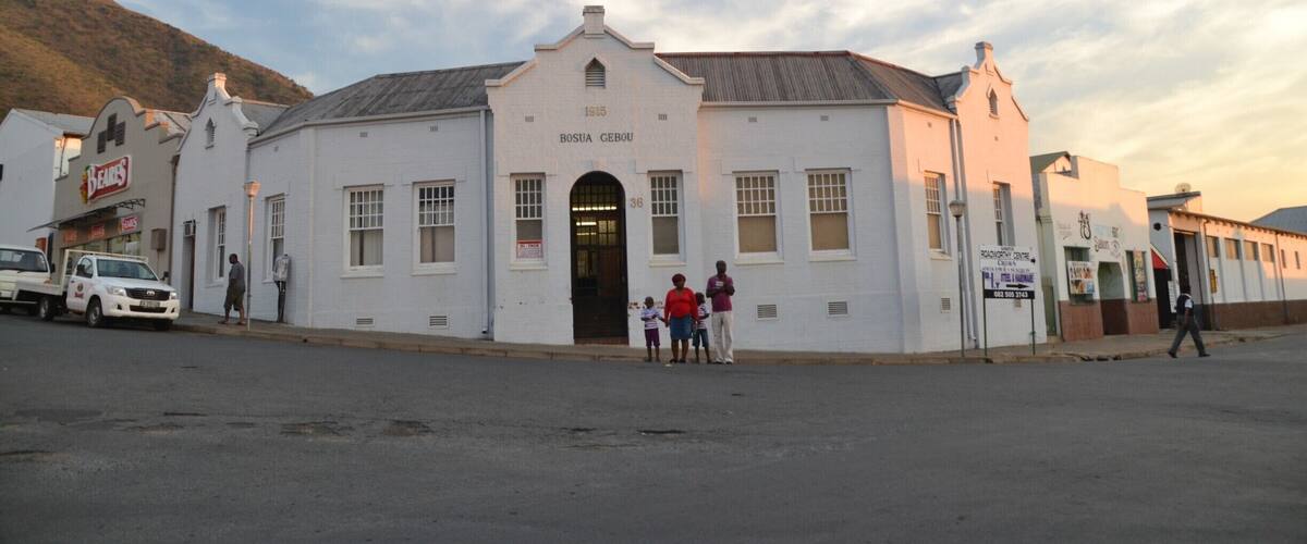 A white building with a sign reading "BOSIA GEBOU" and a few people standing in front.