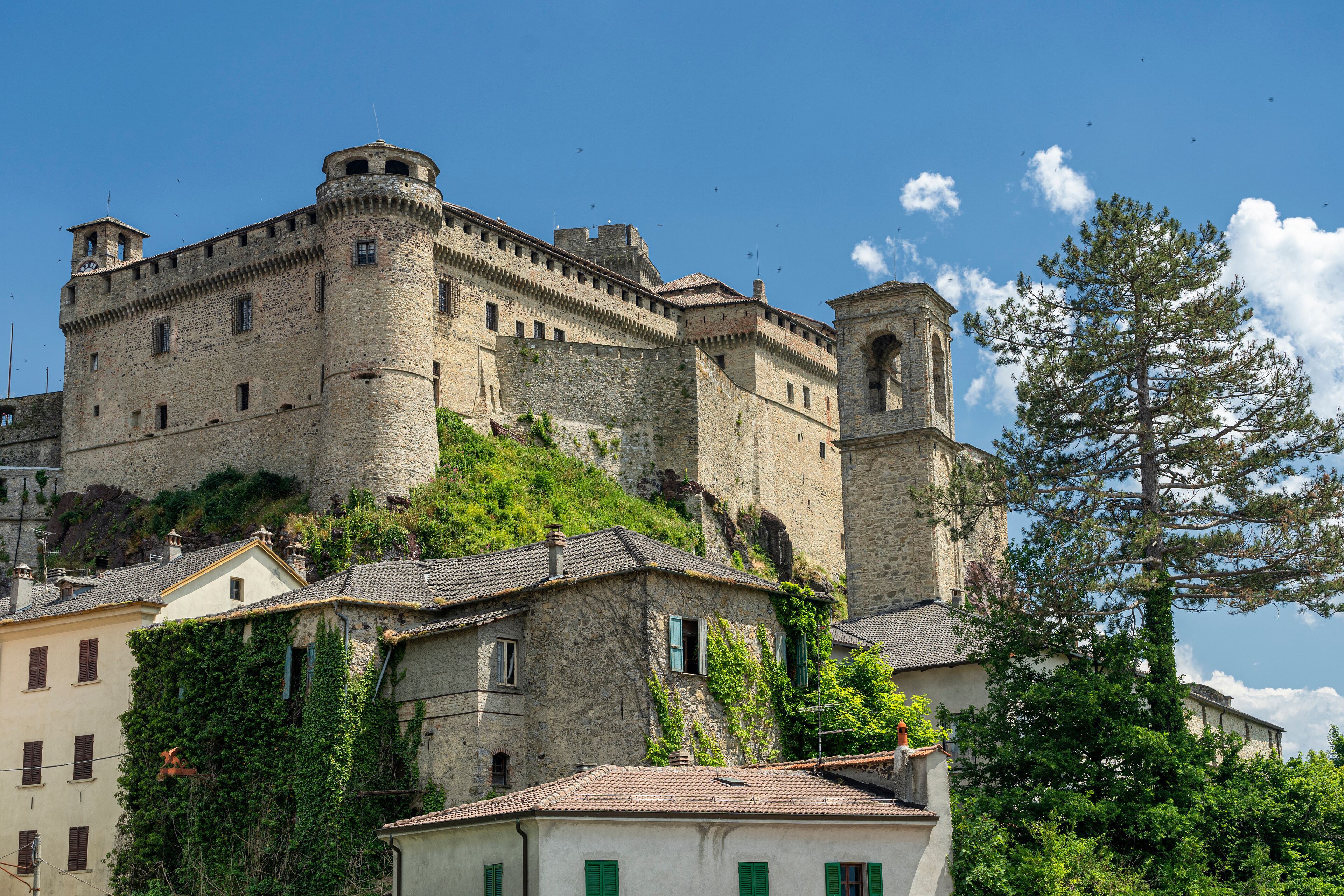 A historic stone castle with a tower, surrounded by lush greenery and traditional buildings.