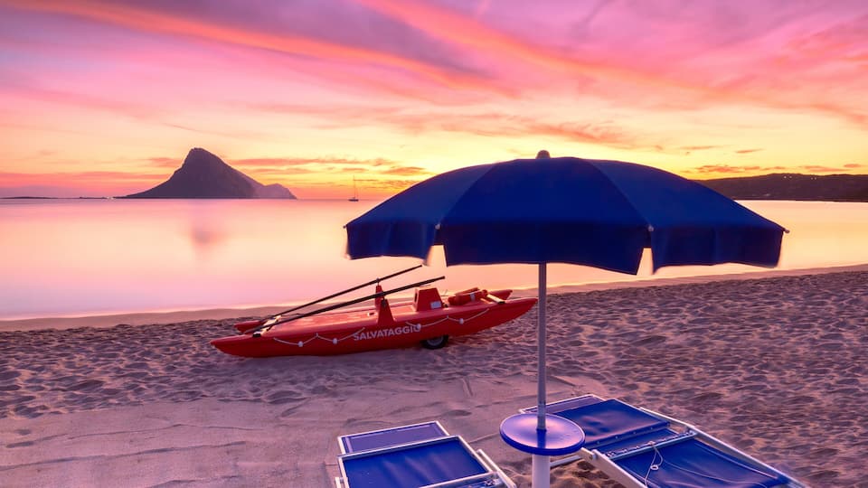 A beach with a red boat, a blue umbrella, and lounge chairs.