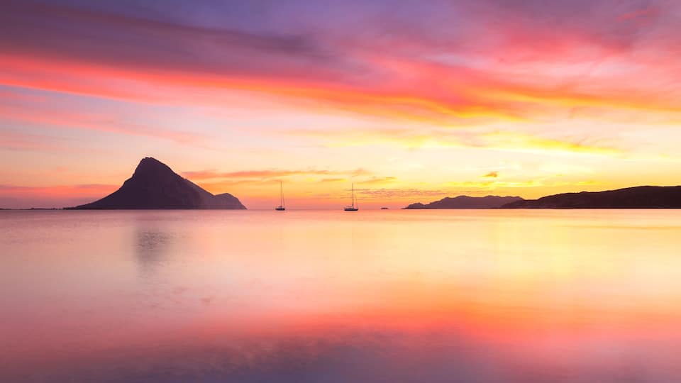A calm sea at sunset with a mountain silhouette and sailboats.
