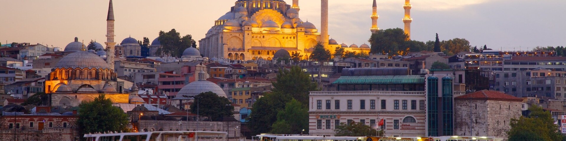 A cityscape with a prominent mosque with multiple domes and minarets, surrounded by urban buildings and a waterfront with boats.