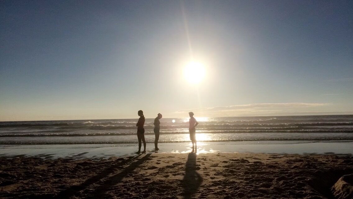 A beach with three people standing near the water's edge, the sun low in the sky, and waves gently crashing onto the shore.