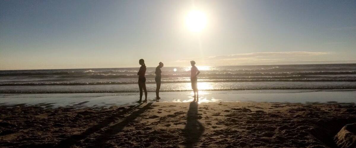 A beach with three people standing near the water's edge, the sun low in the sky, and waves gently crashing onto the shore.