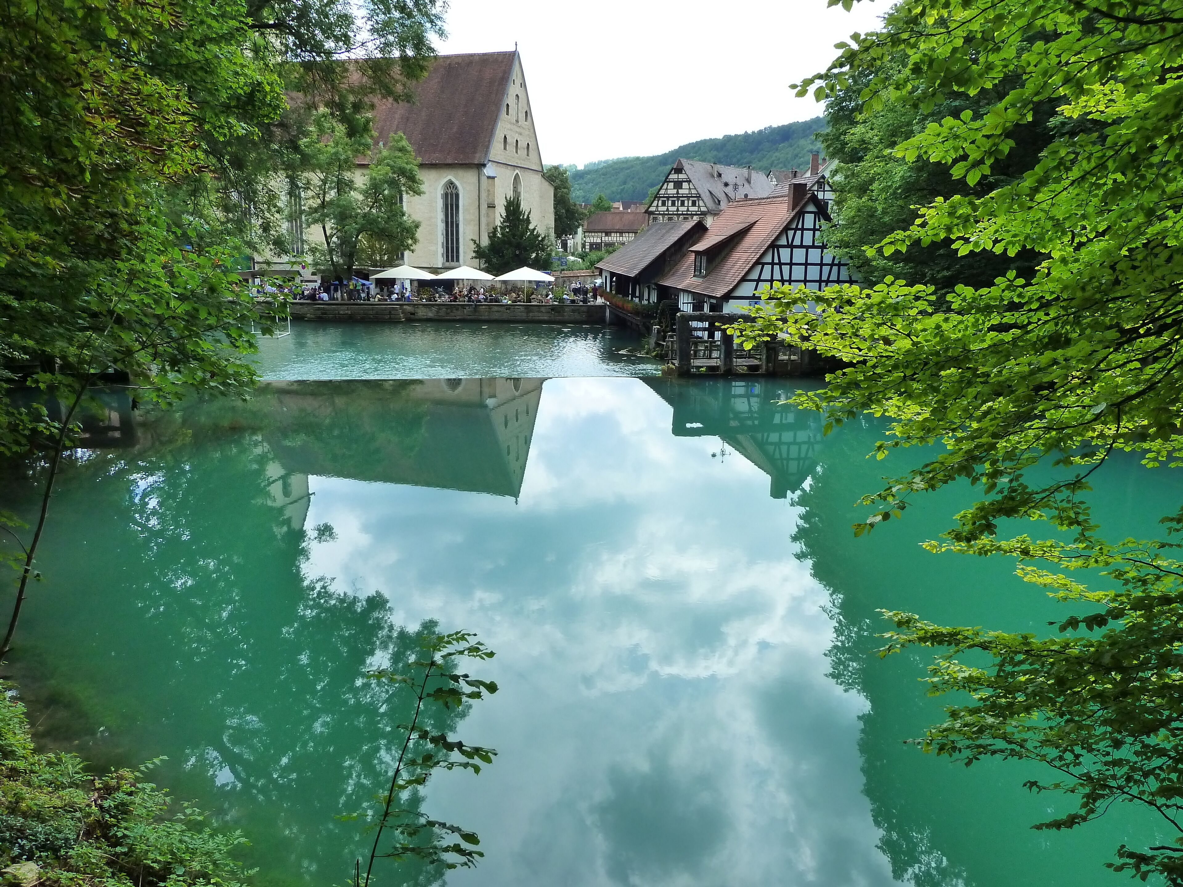 A serene river with a church and traditional buildings on the right, surrounded by lush greenery.