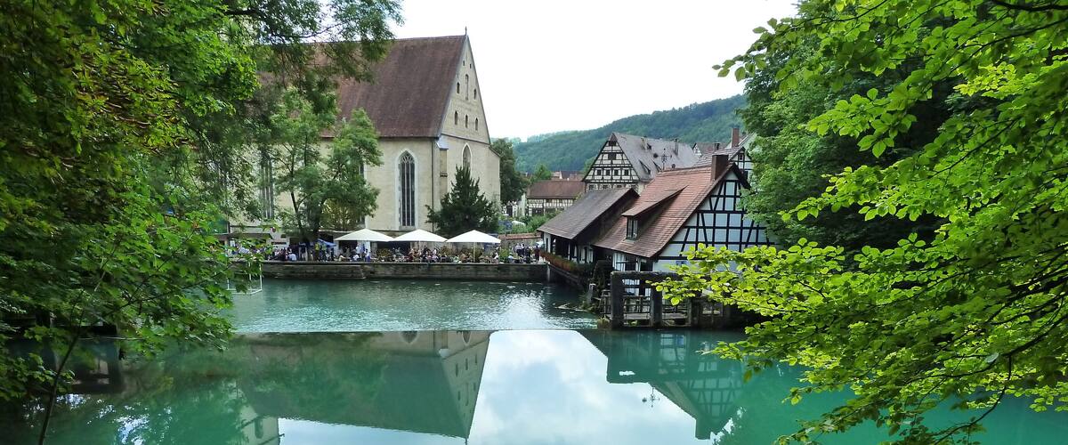 Une rivière paisible, avec une église et des bâtiments traditionnels sur la rive droite, le tout entouré d’une végétation luxuriante.