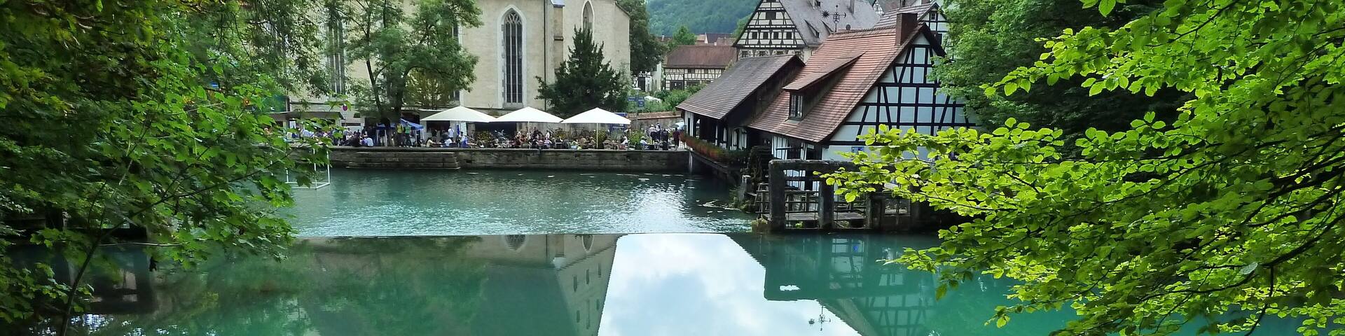 A serene river with a church and traditional buildings on the right, surrounded by lush greenery.