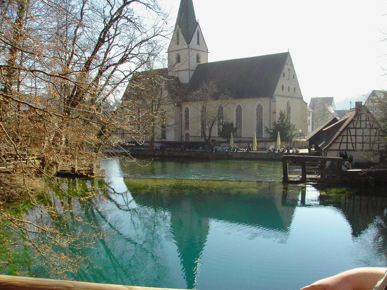 A church with a tall steeple, a calm river, and traditional half-timbered houses.