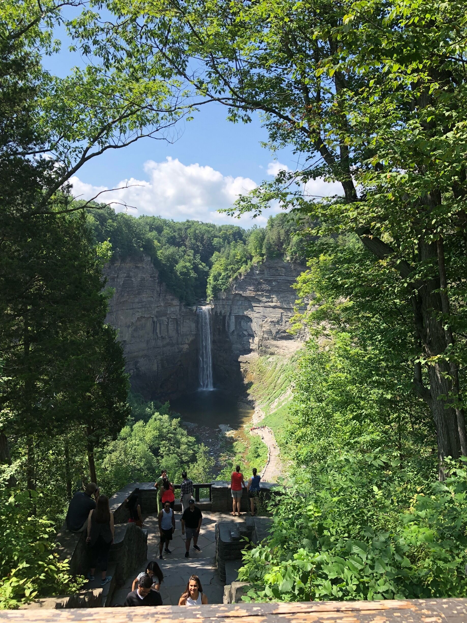 A waterfall cascading down a rocky cliff surrounded by lush greenery and a pathway with visitors.