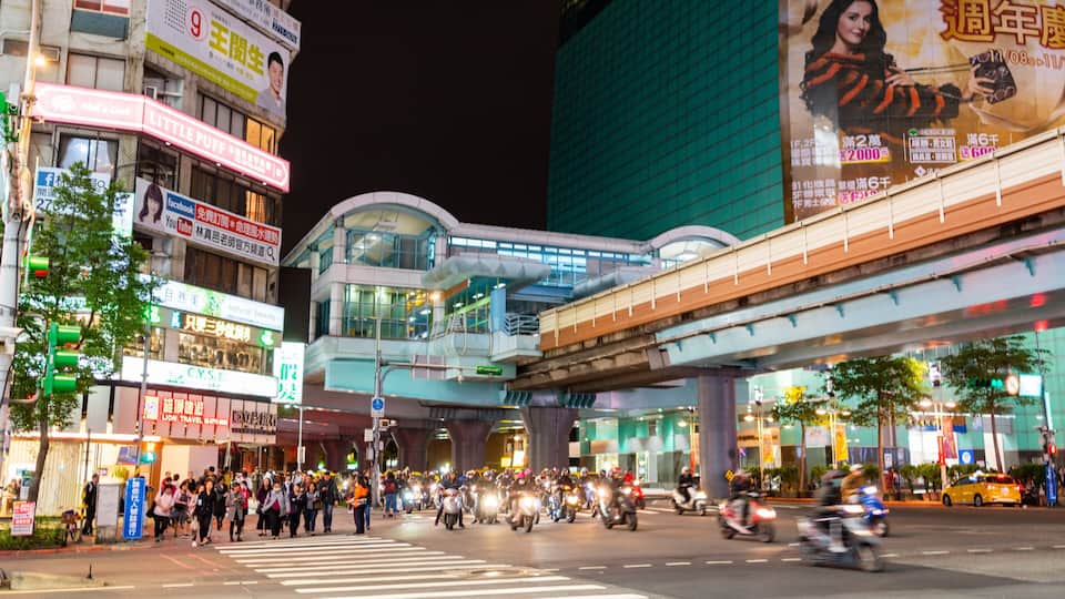 A busy urban street at night with illuminated signs, a pedestrian crossing, and a multi-level overpass.