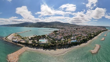 An aerial view of a coastal town with a beach, clear turquoise waters, and a mountainous backdrop.
