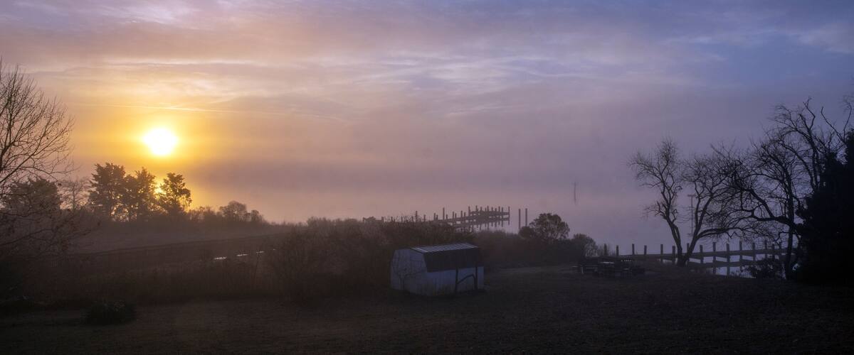 A rural landscape with a barn, trees, and a fence under a sunset sky.