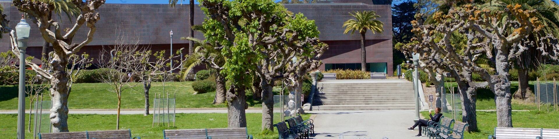 A modern building with a unique angular design, surrounded by a well-maintained park with benches and palm trees.
