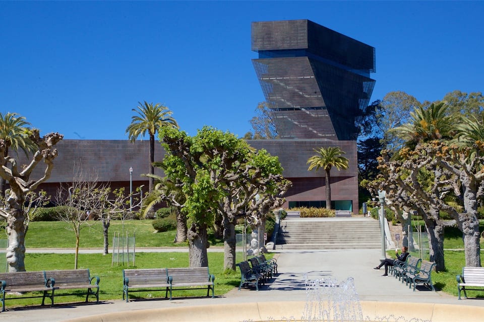 A modern building with a unique angular design, surrounded by a well-maintained park with benches and palm trees.