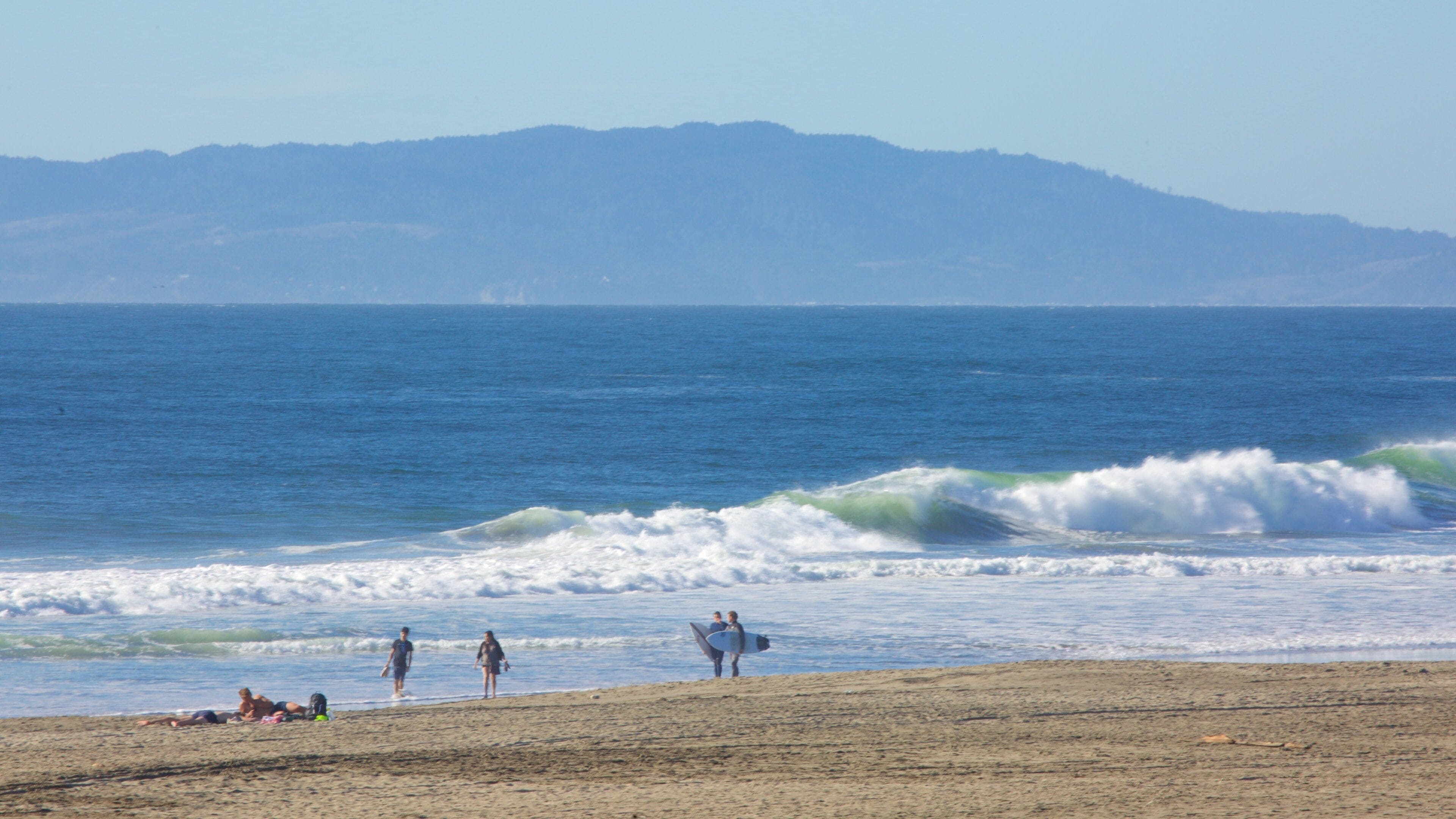 Uma praia com ondas, pessoas e montanhas ao fundo.