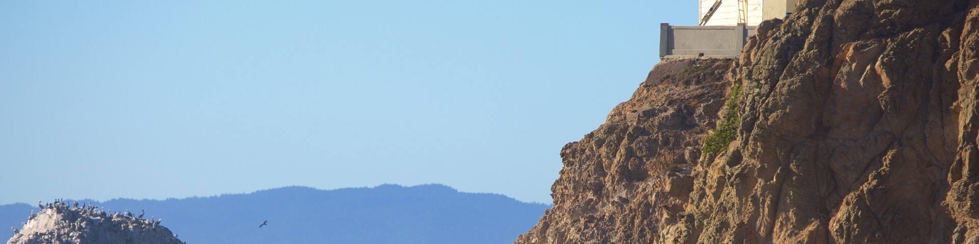 A rocky coastline with a person walking on the beach, a large rock formation, and a distant mountain range.