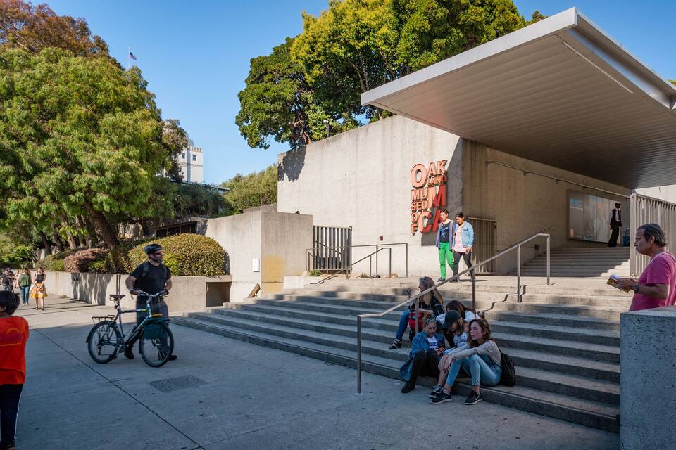 A modern building with a large sign reading "Oakland Museum of California" and a staircase leading to an entrance.