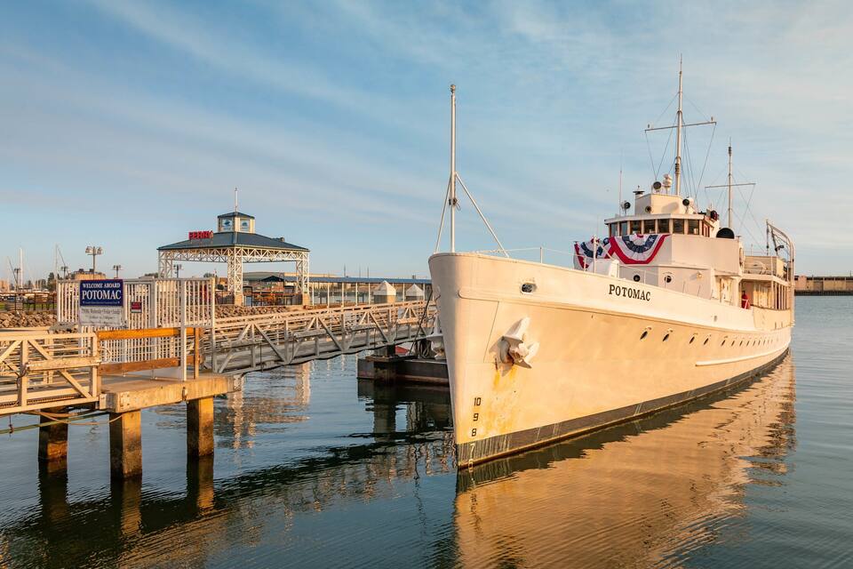 Una grande nave chiamata POTOMAC ormeggiata a un molo con un piccolo edificio su cui è scritto 'BENVENUTI A BORDO DEL POTOMAC'.