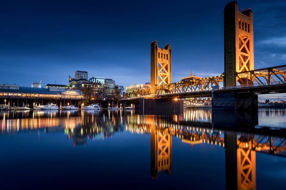 A nighttime view of a bridge with illuminated towers reflecting on the water, a marina with boats, and city buildings in the background.