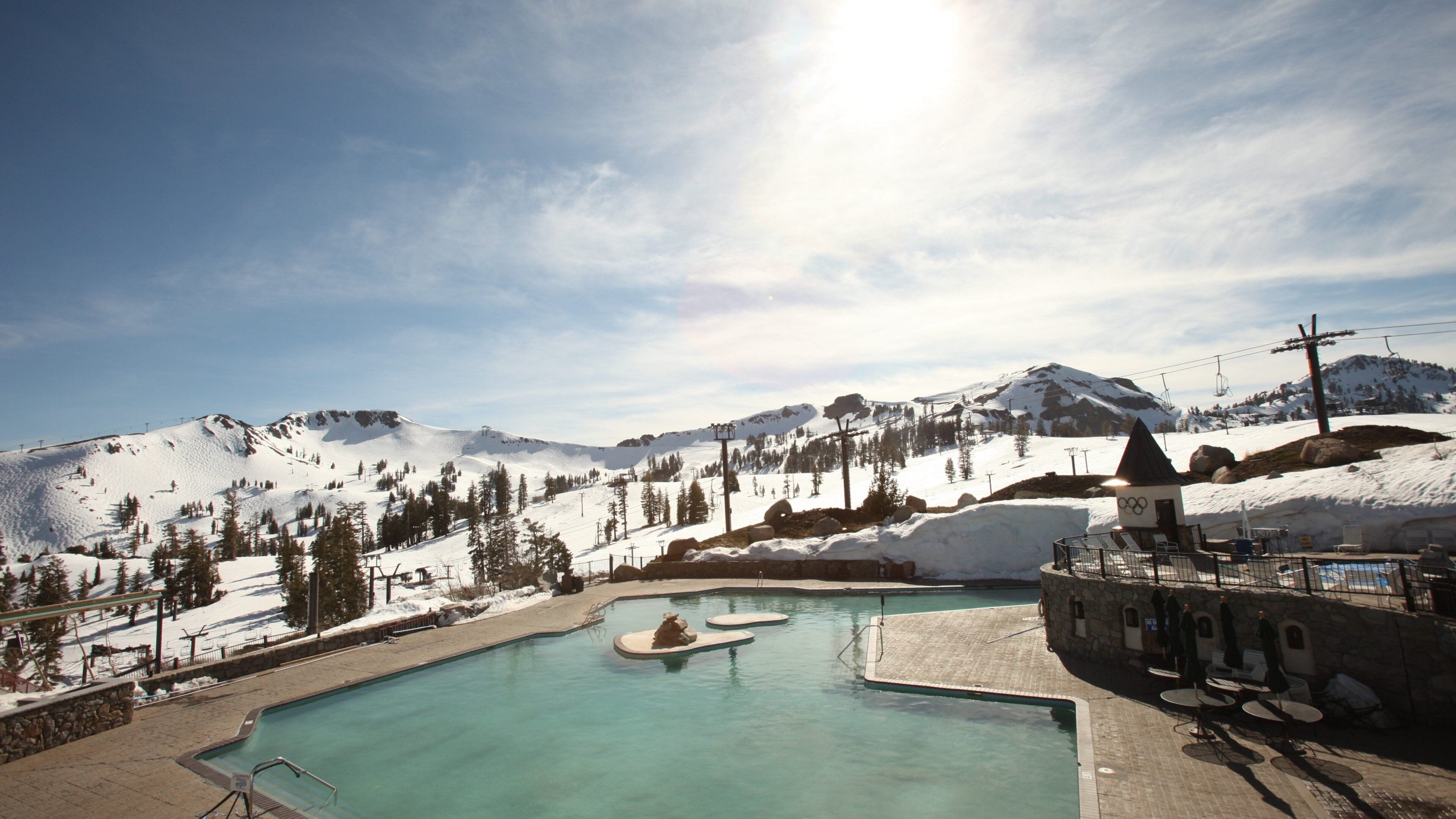 A mountain resort with a swimming pool surrounded by snow-covered mountains and a ski lift.