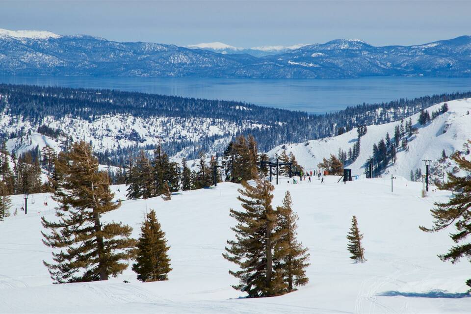 A snowy mountain landscape with a ski lift, trees, and a lake in the background.