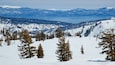 A snowy mountain landscape with a ski lift, trees, and a lake in the background.