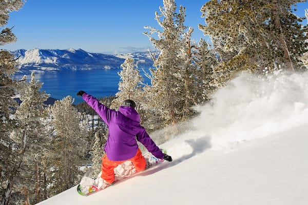 Ein Snowboarder mit lila Jacke und orangefarbenen Hosen an einem verschneiten Hang, im Hintergrund ein Berg und ein See.