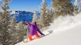 A snowboarder in a purple jacket and orange pants on a snowy slope with a mountain and lake in the background.