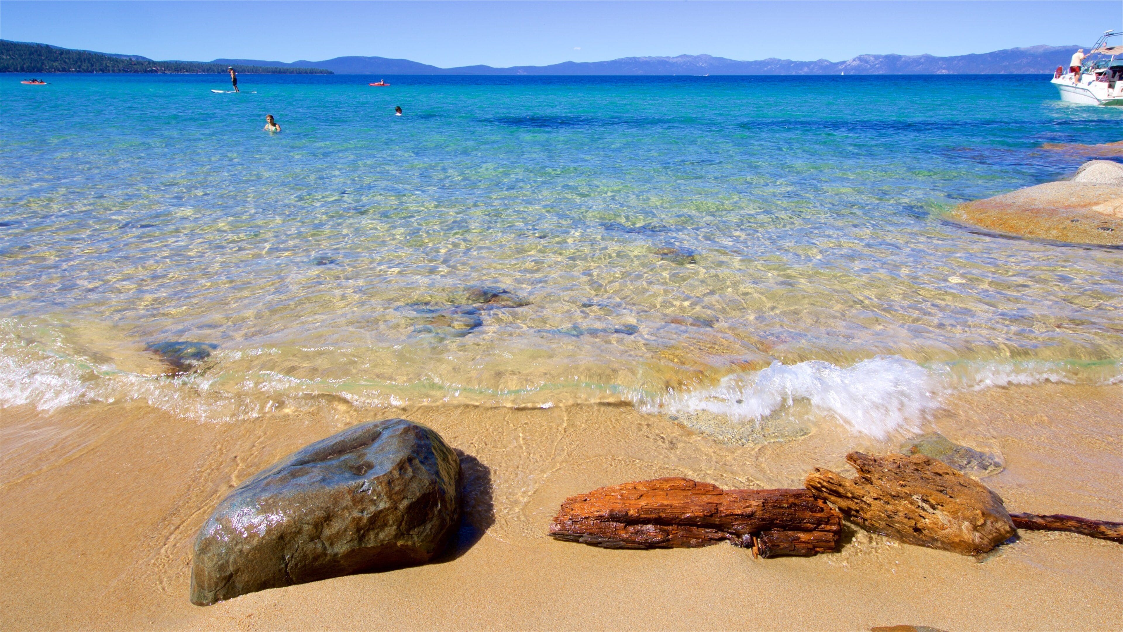 A clear turquoise sea with a sandy beach, large rocks, and distant mountains.