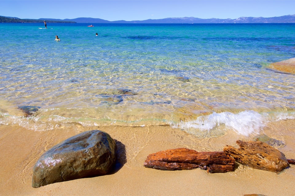 A clear turquoise sea with a sandy beach, large rocks, and distant mountains.