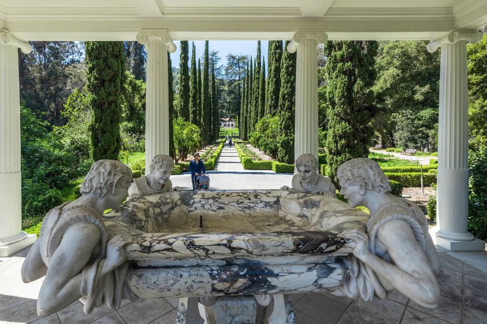 Um jardim clássico com uma fonte de mármore, árvores altas e um caminho que leva a um edifício distante.