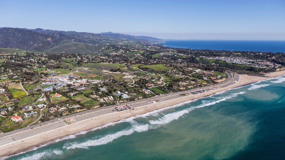 An aerial view of a coastal town with a beach, roads, and surrounding hills.