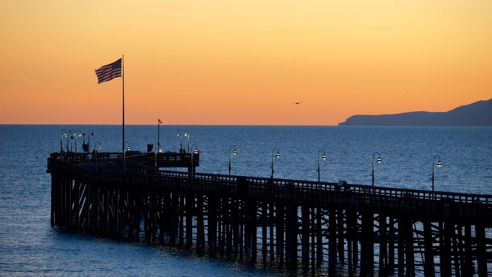 A pier extending into the water with a flag on top, street lamps, and a sunset in the background.