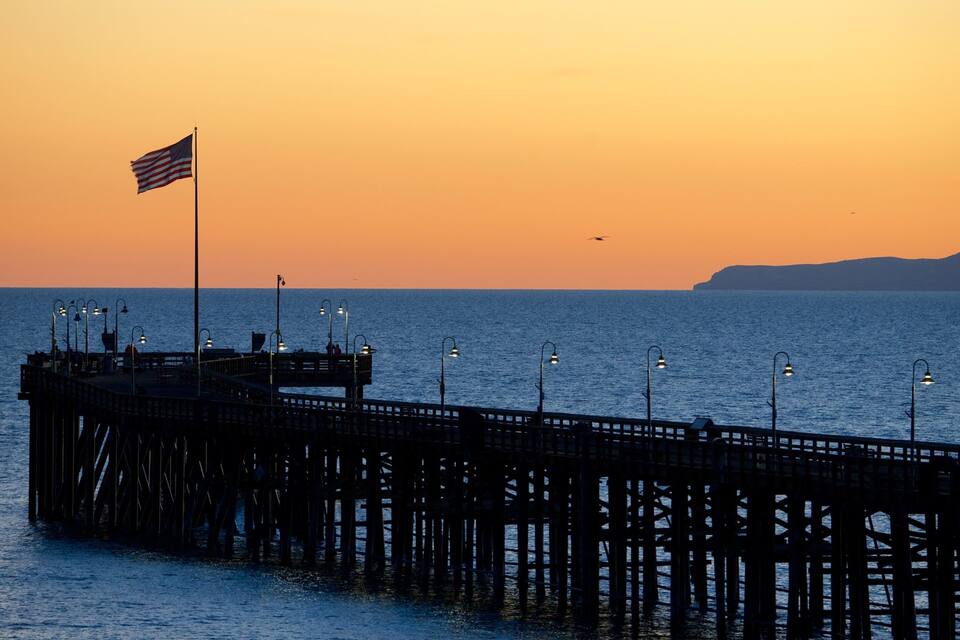 A pier extending into the water with a flag on top, street lamps, and a sunset in the background.
