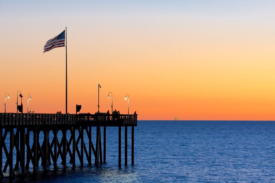 Een steiger die zich uitstrekt over het water, met een Amerikaanse vlag, lantaarns en mensen die op het dek zitten tijdens de zonsondergang.