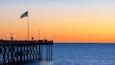 A pier extending into the water with an American flag, street lamps, and people sitting on the deck during sunset.