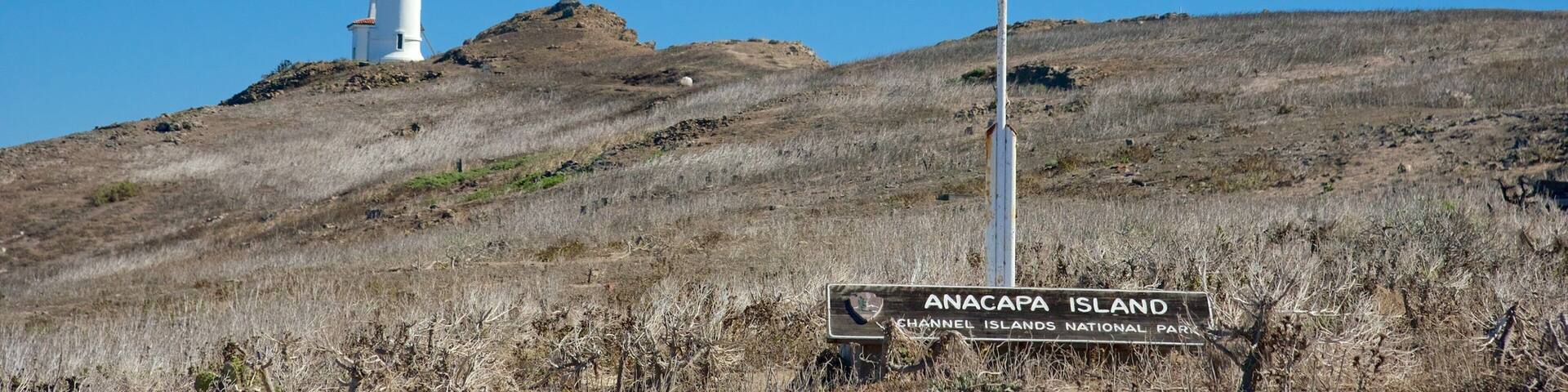 A flagpole with an American flag, a lighthouse, and a sign for Anacapa Island in the background.