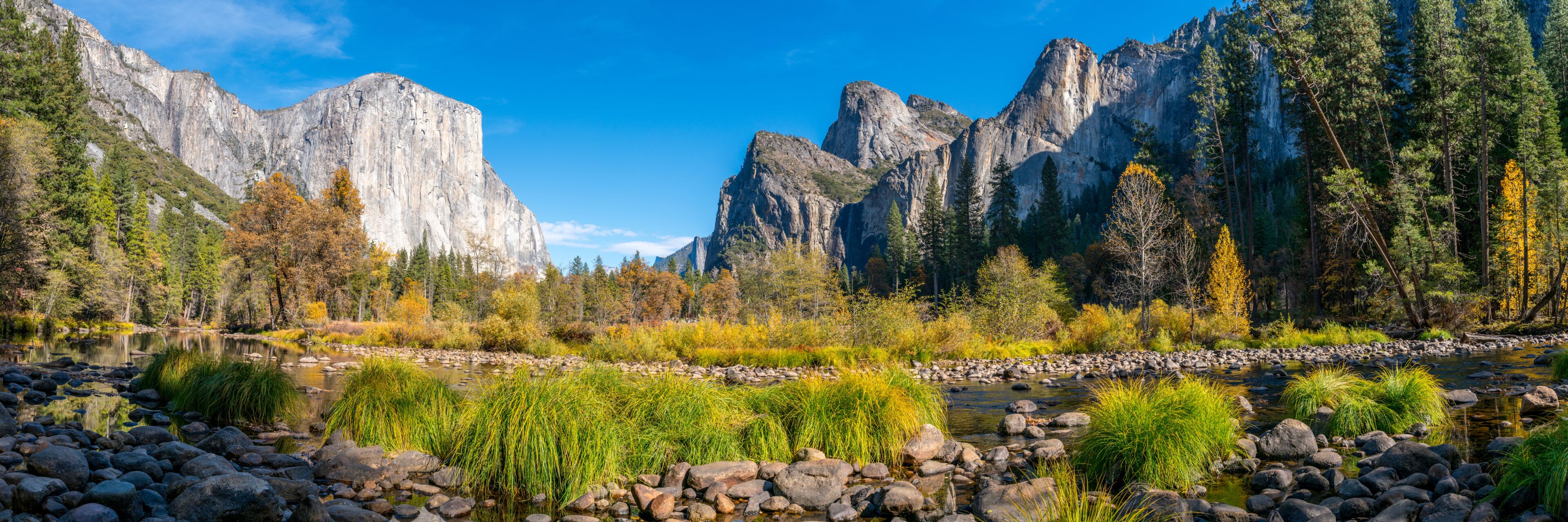 A river with rocks and vegetation, surrounded by mountains and autumn-colored trees.