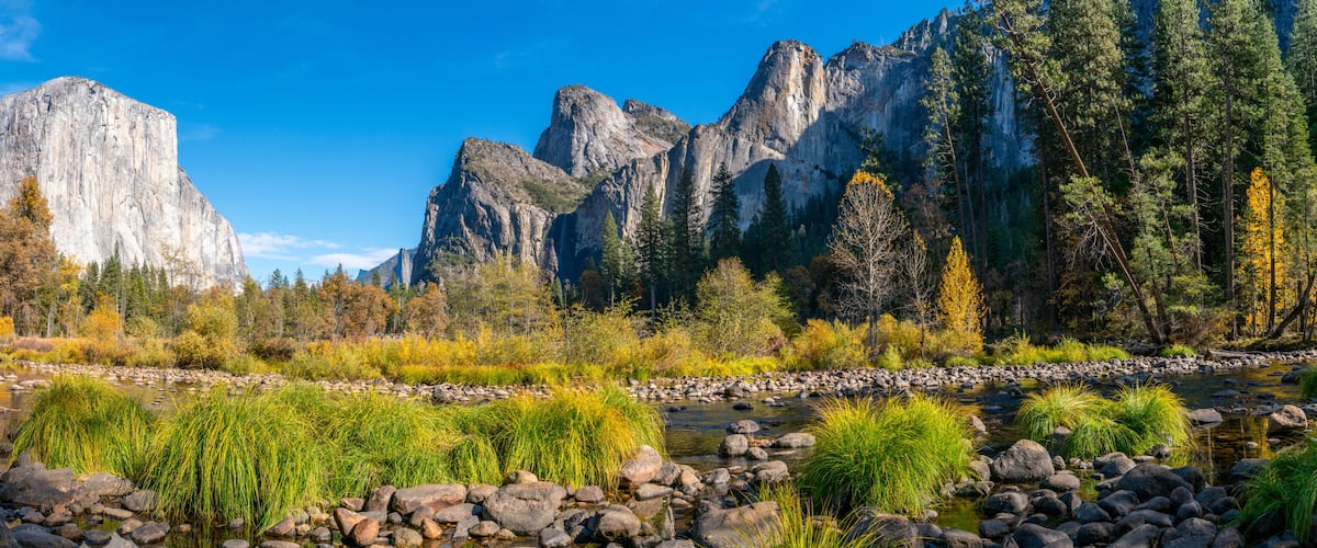 A river with rocks and vegetation, surrounded by mountains and autumn-colored trees.