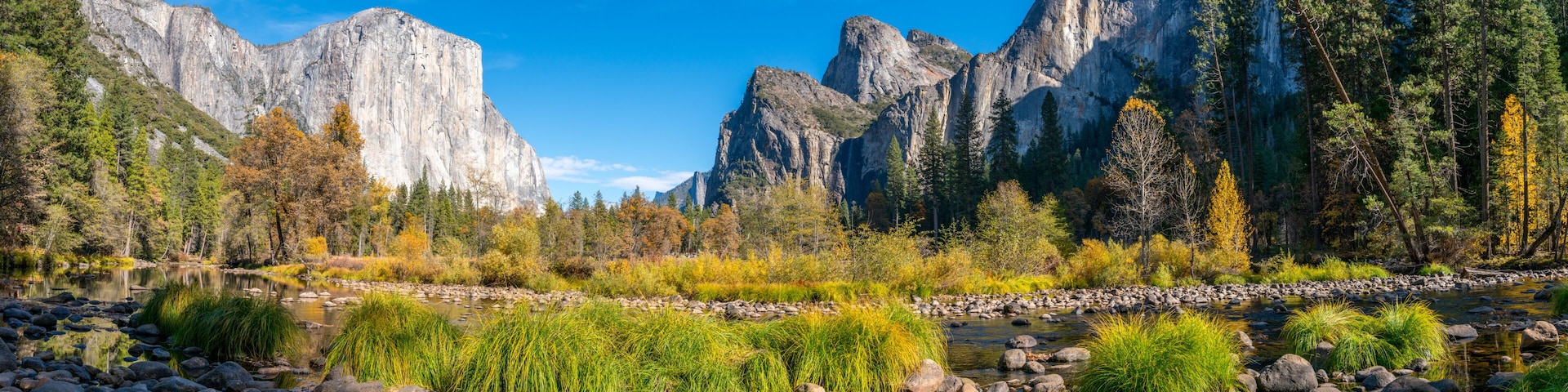A river with rocks and vegetation, surrounded by mountains and autumn-colored trees.