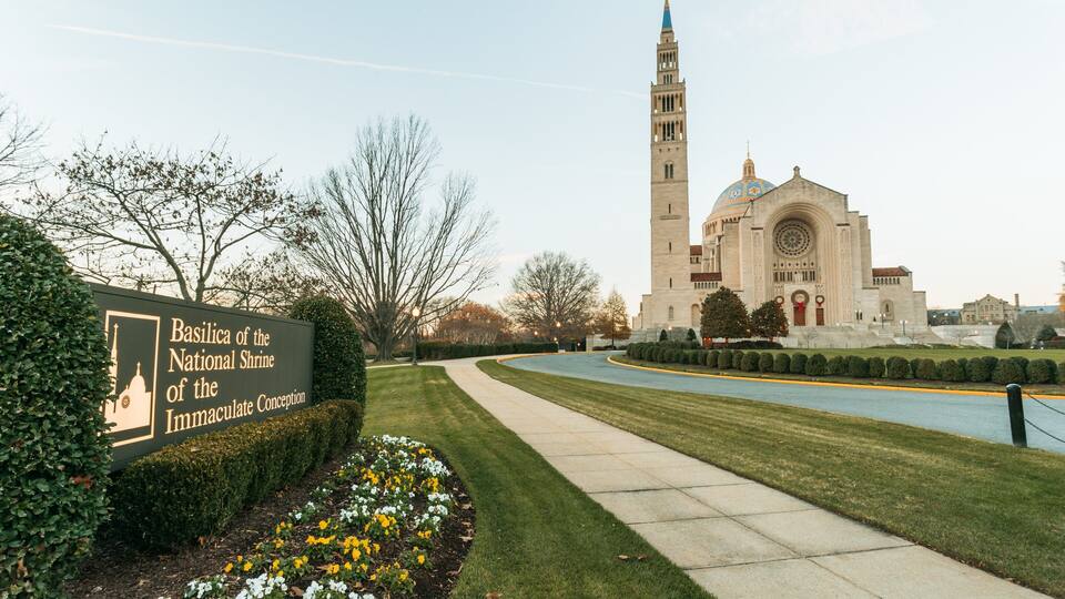 A sign for Basilica of the National Shrine of the Immaculate Conception, a large church with a tall tower and dome, surrounded by well-maintained lawns and trees.