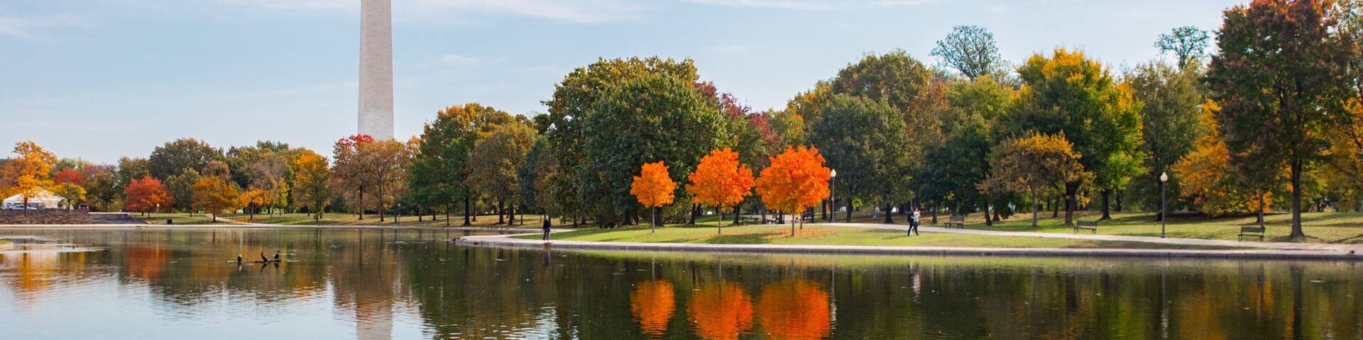 A lake with autumn trees reflecting in the water, a tall monument in the background, and a clear sky.