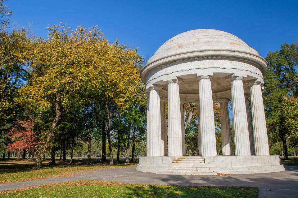 A classical white marble gazebo with columns, surrounded by trees with autumn foliage.