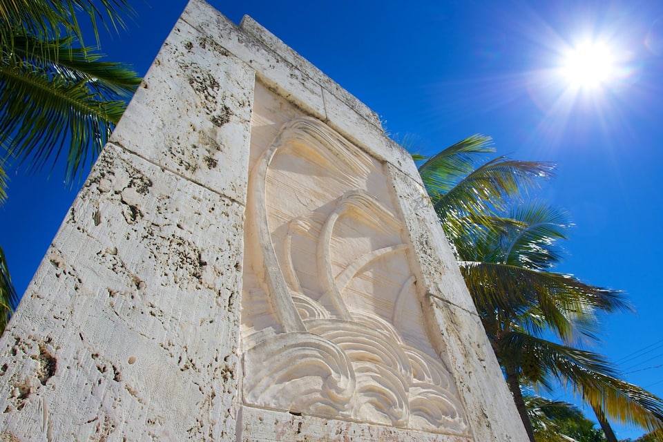 A weathered stone monument with a carved wave design, surrounded by palm trees and under a clear blue sky.
