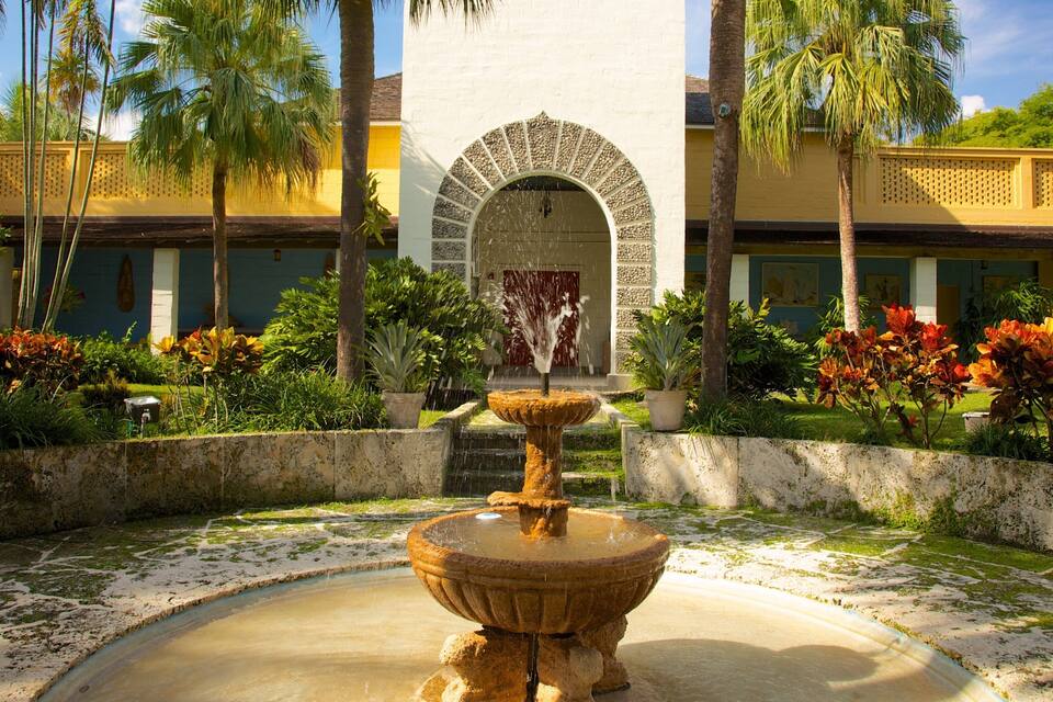 A courtyard with a central fountain, palm trees, and a building with a distinctive arched entrance.