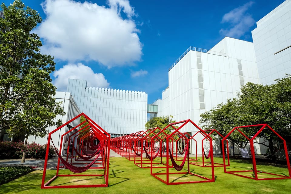 Un complejo moderno de edificios con fachadas de vidrio reflectante, rodeado de un césped verde y esculturas geométricas rojas.